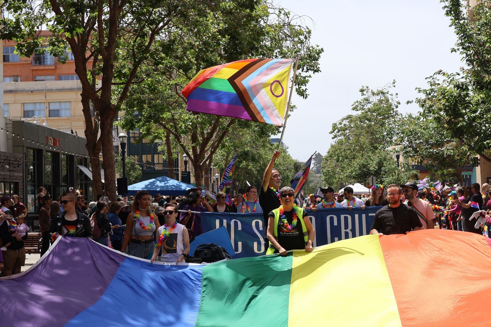 The Santa Cruz Pride parade, marchers carrying a giant rainbow flag down the street under a Progress Pride flag.
